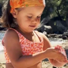 Petite fille assise dans le sable, dos à la mer, appliquant de la crème solaire bébé bio sur son corps.