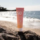 Tube de crème solaire visage vegan posé sur le sable, avec la plage et le soleil du sud de la France en arrière-plan.