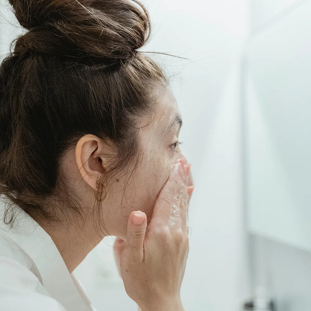 Jeune femme nettoyant son visage avec un gel purifiant anti-imperfections bio devant son miroir